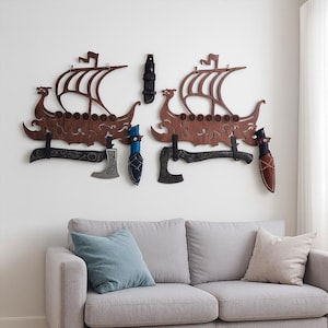 Puede incluir: Dos colgantes de pared con forma de barco vikingo de madera marrón, cada uno con un hacha y un cuchillo decorativos. Las hachas y los cuchillos son negros y plateados, con fundas azules y marrones. Los barcos están montados en una pared blanca.