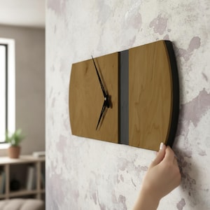 May include: A rectangular wooden wall clock with a light brown face, black hands, and a dark gray vertical stripe. The clock has black curved edges and is mounted on a wall.