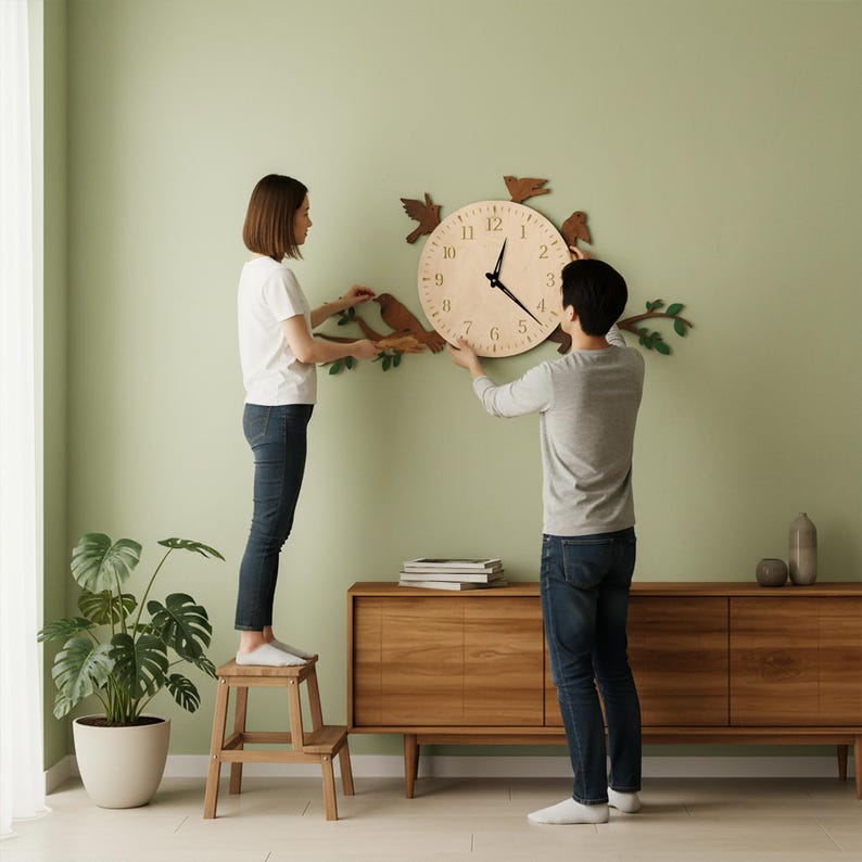 May include: A wooden wall clock with a bird and branch design. The clock face is light-colored with black hands and numerals. Decorative birds and branches are attached around the clock. The clock is being installed on a light green wall.