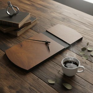May include: A wooden wall clock with black hands, a dark brown face, and a light brown top section. A cup of coffee sits nearby on a wooden table, along with a stack of books and a silver object.