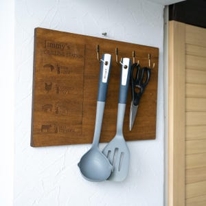 May include: A wooden grilling station with a thermometer chart for different types of meat. The station has hooks for hanging kitchen utensils, including a grey ladle and spatula, and a pair of black kitchen shears.