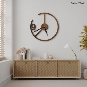 May include: Teak-colored wooden wall clock with a unique open design. The clock features a circular frame with the number 9 and black clock hands. The clock is mounted on a white wall above a woven cabinet.