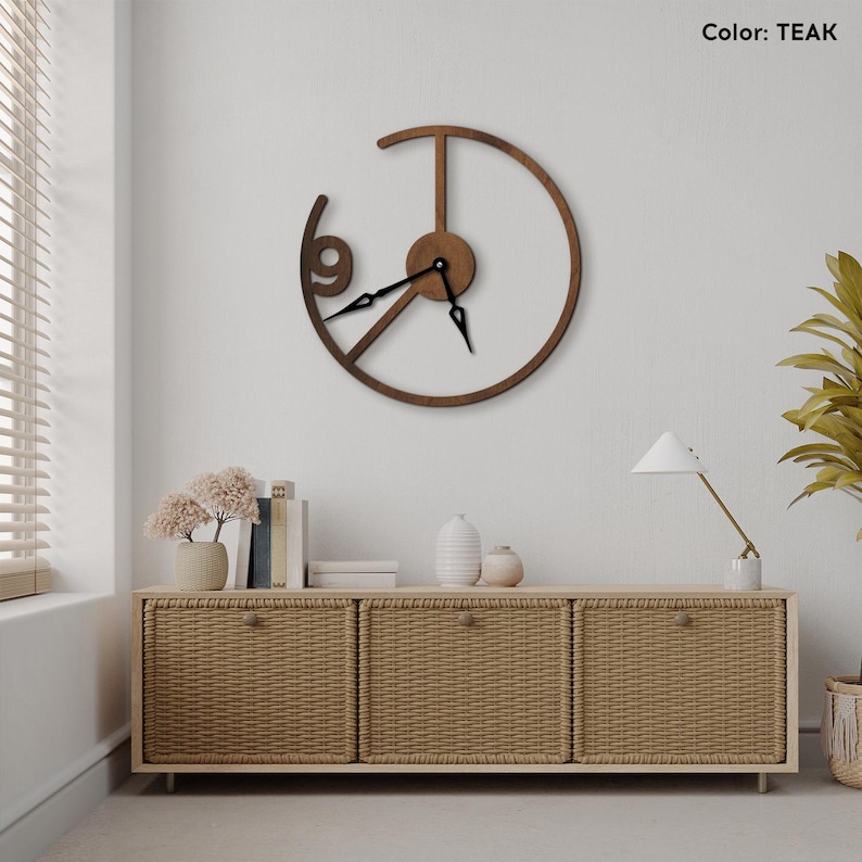 May include: Teak-colored wooden wall clock with a unique open design. The clock features a circular frame with the number 9 and black clock hands. The clock is mounted on a white wall above a woven cabinet.
