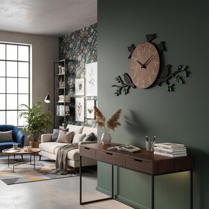 May include: A brown wooden wall clock with a bird and branch design. The clock has a round face with black hands and numbers. The birds and leaves are black and green. The clock is mounted on a dark green wall in a living room.