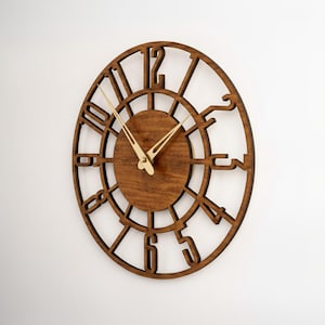 Puede incluir: Un reloj de pared de madera con acabado marr&oacute;n oscuro. El reloj presenta un dise&ntilde;o circular con n&uacute;meros recortados y una esfera central. Las manecillas doradas indican la hora. El reloj est&aacute; montado en una pared blanca.