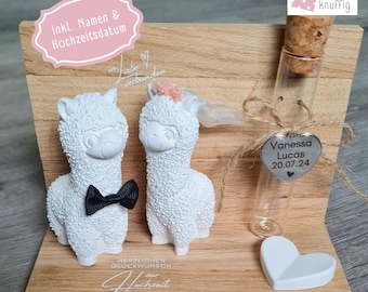 May include: A wooden wedding decoration with two white llama figurines, one with a black bow tie and the other with a pink floral headpiece. A glass vial with a heart-shaped tag and the names "Vanessa Lucas 20.07.24" is also included.
