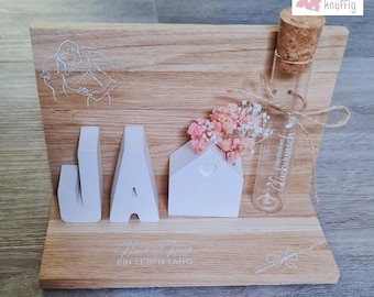 Peut inclure: Une pancarte en bois avec le mot "JA" en lettres blanches, une petite maison blanche avec une découpe en forme de cœur et un flacon en verre avec des fleurs séchées. La pancarte porte également le texte "Hand in Hand Ein Leben Lang" et "Mit Liebe für Dich in Deutschland hergestellt."