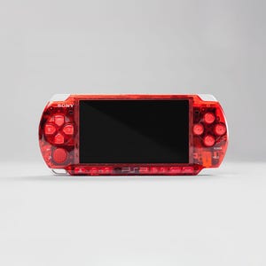 Pode incluir: Um console de jogos portátil PSP vermelho da Sony. O dispositivo possui uma caixa vermelha transparente com uma tela preta e botões vermelhos. O logotipo da Sony é visível no canto superior esquerdo.