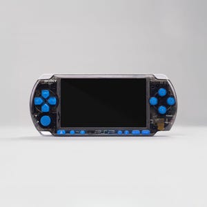 May include: A black and clear handheld gaming console with blue buttons. The console has a black screen and the word "SONY" is visible at the top. The console has a directional pad, action buttons, and other control buttons.