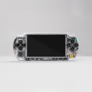 Puede incluir: Una consola de juegos portátil transparente con componentes internos visibles. El dispositivo presenta una pantalla negra, un panel direccional, botones de acción y varios puertos. La carcasa transparente permite ver los circuitos y componentes internos.