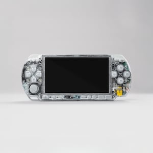 Pode incluir: Uma consola de jogos portátil transparente, revelando os seus componentes internos. O dispositivo possui um ecrã preto, um painel direcional e botões de ação, tudo numa caixa de plástico transparente. O design mostra os circuitos e componentes internos.
