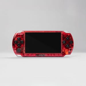 Pode incluir: Um console de jogos portátil Sony PSP vermelho. O console tem uma caixa vermelha transparente, com botões pretos e uma tela preta. O PSP tem um D-pad, botões de ação e um joystick. O logotipo da PSP é visível na parte inferior.
