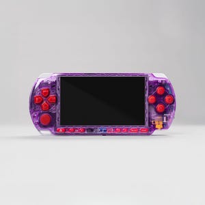 Könnte beinhalten: Eine tragbare Spielkonsole mit einem durchscheinenden violetten Kunststoffgehäuse und leuchtend roten Tasten. Das Gerät verfügt über einen zentralen rechteckigen Bildschirm, ein Steuerkreuz links und vier Aktionstasten rechts. Das Sony-Logo ist sichtbar.