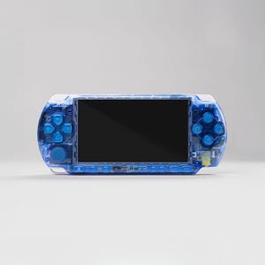 Könnte beinhalten: Eine transparente blaue Handheld-Spielkonsole mit schwarzem Bildschirm. Die Konsole hat blaue Tasten und eine weiße Oberseite. Der Markenname ist sichtbar. Das Design ist schlank und modern.