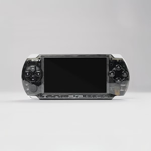 Könnte beinhalten: Eine schwarze und transparente Sony PSP-Handheld-Spielkonsole. Die Konsole hat einen schwarzen Bildschirm, ein Steuerkreuz und Aktionstasten. Die PSP hat eine weiße Oberseite und ist vor einem weißen Hintergrund platziert.