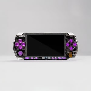 May include: A handheld gaming console with a transparent black casing and vibrant purple buttons. The device features a central screen, a directional pad on the left, four action buttons on the right, and the Sony logo printed above the screen.