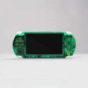 May include: A handheld gaming console with a translucent emerald green plastic shell. The device features a central black screen, a directional pad on the left, four action buttons on the right, and the Sony and PSP logos printed on the front.