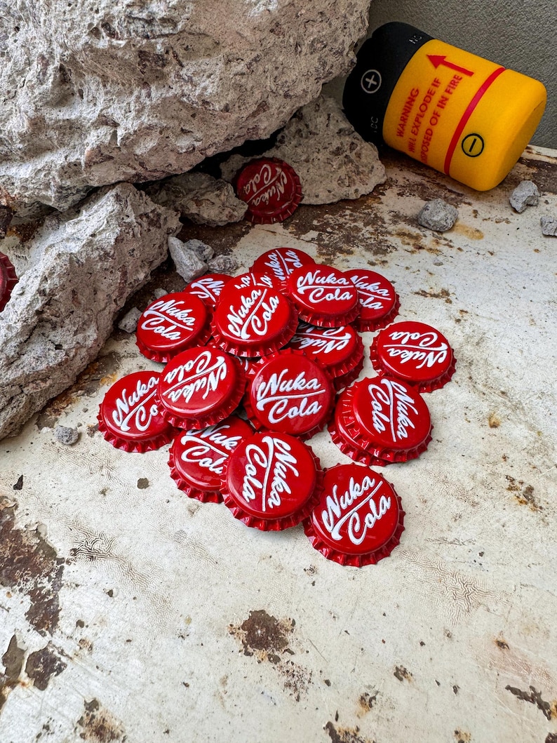 May include: A pile of red bottle caps with the words "Nuka Cola" printed in white. The caps are on a rusty metal surface.