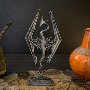 Könnte beinhalten: Eine metallisch grau-bronze Skulptur des Skyrim-Logos mit einem Drachendesign. Die Skulptur steht auf einem kleinen Sockel, vor dem Hintergrund einer Karte und Münzen. Im Hintergrund ist eine orangefarbene Flasche zu sehen.