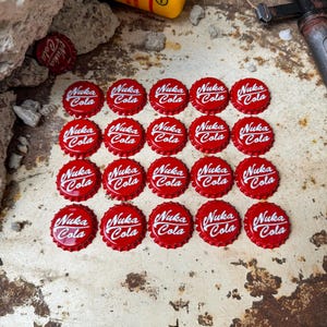 May include: Twenty red bottle caps with the words "Nuka Cola" in white lettering. The bottle caps are arranged in a grid pattern on a rusty metal surface.