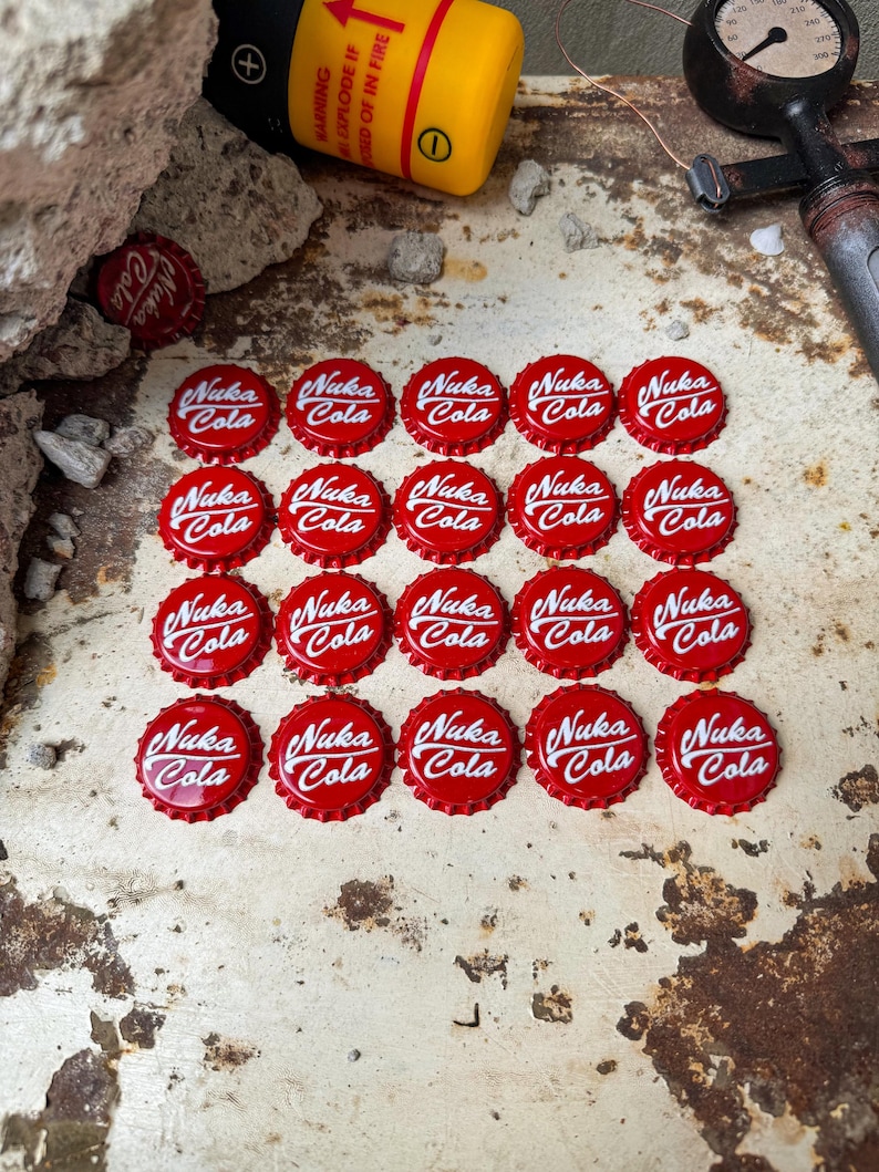 May include: Twenty red bottle caps with the words "Nuka Cola" in white lettering. The bottle caps are arranged in a grid pattern on a rusty metal surface.