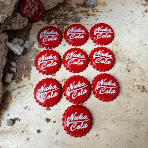 May include: Twelve red bottle caps with the white text "Nuka Cola" on each. The bottle caps are arranged in a pyramid shape on a white surface.