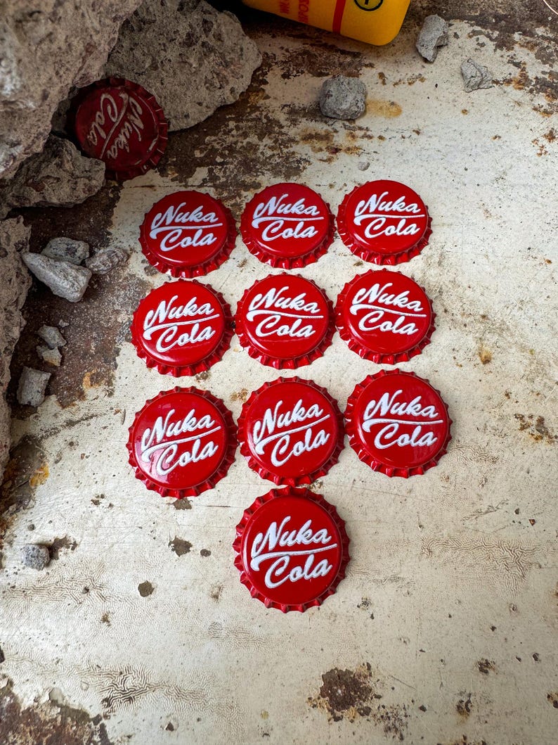 May include: Twelve red bottle caps with the white text "Nuka Cola" on each. The bottle caps are arranged in a pyramid shape on a white surface.