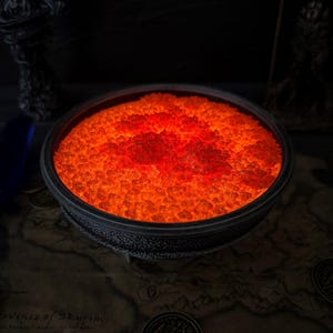 May include: A circular, dark bowl filled with glowing orange crystals, reminiscent of hot embers. The bowl rests on a map of Skyrim, with two decorative statues in the background. The image evokes a fantasy or gaming theme.
