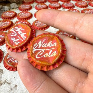 Może przedstawiać: Dwa czerwone i zardzewiałe korki do butelek z białym napisem "Nuka Cola".
