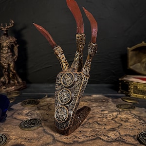 Op de afbeelding: Een gedetailleerde fantasieklauwprop met drie lange, rood getipte klauwen. De handvormige basis is bruin met gouden accenten en drie cirkelvormige emblemen. De prop staat op een kaart met gouden munten verspreid.