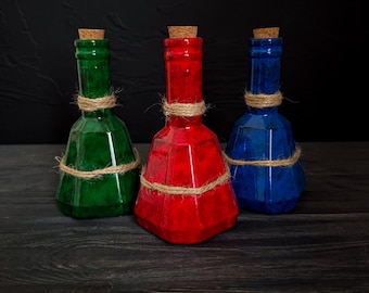 May include: Three colorful glass bottles with cork stoppers. The bottles are green, red, and blue, each wrapped with twine around the neck. The bottles have a faceted design and sit on a dark wooden surface.