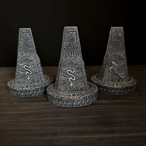 May include: Three gray stone obelisks with carved spiral and serpent designs. Each obelisk has a circular base and a tapered shape, with a textured, weathered appearance. The background is dark.