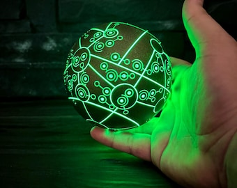 Op de afbeelding: Een bolvormig object met een donker, getextureerd oppervlak met ingewikkelde, oplichtende groene geometrische patronen van onderling verbonden cirkels en lijnen. De bol straalt van binnenuit een helder groen licht uit.