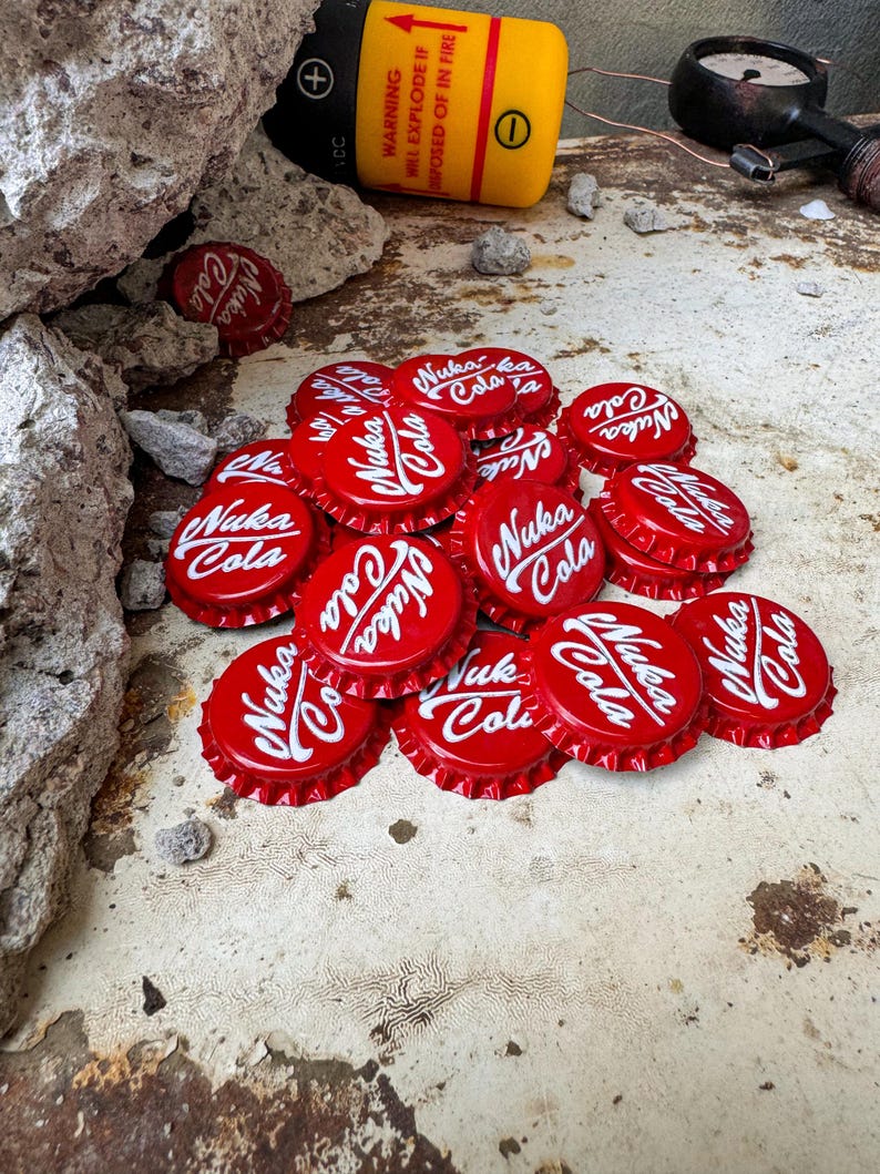 May include: A pile of red bottle caps with the white text "Nuka Cola" on them. The caps are scattered on a rusty metal surface.