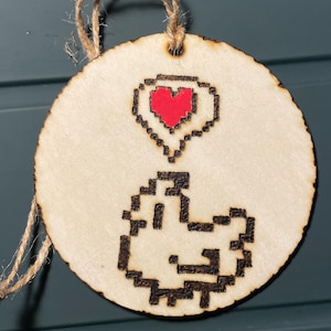 Peut inclure: Une décoration en bois avec une image pixelisée d'un fantôme tenant un cœur rouge. La décoration est ornée d'une ficelle en ficelle brune pour l'accrocher.