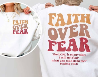 May include: White sweatshirt with the words "FAITH OVER FEAR" in orange, brown, and red. The back of the sweatshirt has the same text, along with a Bible verse. The sweatshirt is a casual, comfortable style.