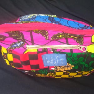 May include: A colorful zippered pouch with a pink, black, yellow, and blue patterned fabric. The pouch has a gold zipper and a white label with the word "Waft" written in black.