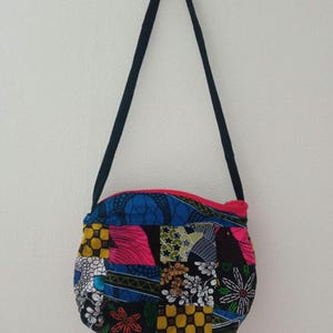May include: A small, round, patchwork purse with a black strap. The purse is made of colorful fabric with a variety of patterns, including floral, geometric, and abstract designs. The purse has a pink zipper.