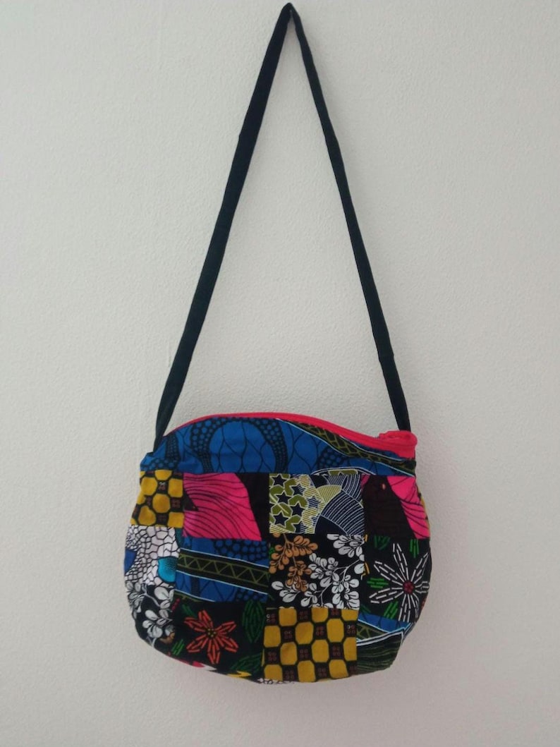 May include: A small, round, patchwork purse with a black strap. The purse is made of colorful fabric with a variety of patterns, including floral, geometric, and abstract designs. The purse has a pink zipper.