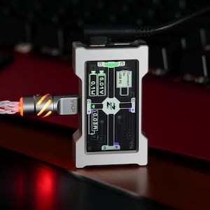 May include: A silver electronic device with a screen showing technical data, connected to a braided white and orange cable. A black cable is plugged into the top. The background is a dark red surface and a keyboard.