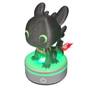 May include: A black dragon figurine with green eyes and an orange harness, mounted on a silver base with a green glowing light. The base has a power button. A small red flag is attached to the dragon's tail.