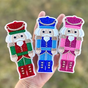 May include: Three colorful nutcracker ornaments in red, blue, and pink, each with glitter accents. The ornaments have white hair and mustaches, and are held in a hand against a blurred background. The ornaments are Christmas themed.