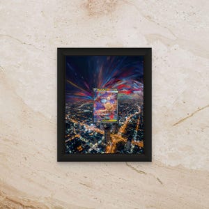 Puede incluir: Una obra de arte enmarcada con un diseño abstracto vibrante de rayas de colores sobre un fondo oscuro. La obra de arte se encuentra sobre un paisaje urbano nocturno, con una carta coleccionable en el centro. El marco es negro.