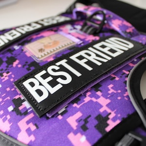 May include: A purple and black digital camouflage dog harness featuring a rectangular black patch with the white text BEST FRIEND. The harness includes a top handle, a metal D-ring for leash attachment, and a clear plastic ID card holder slot.