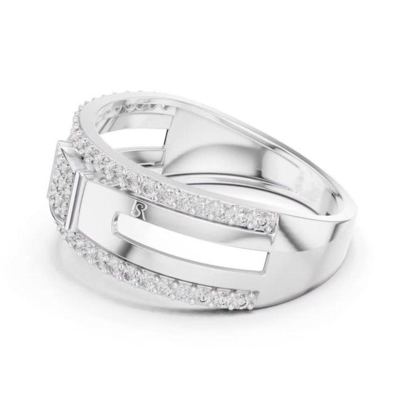 May include: A silver ring with a rectangular center and a band lined with small, clear stones. The ring has a modern design with a polished finish and a small, stylized letter "R" on the side.