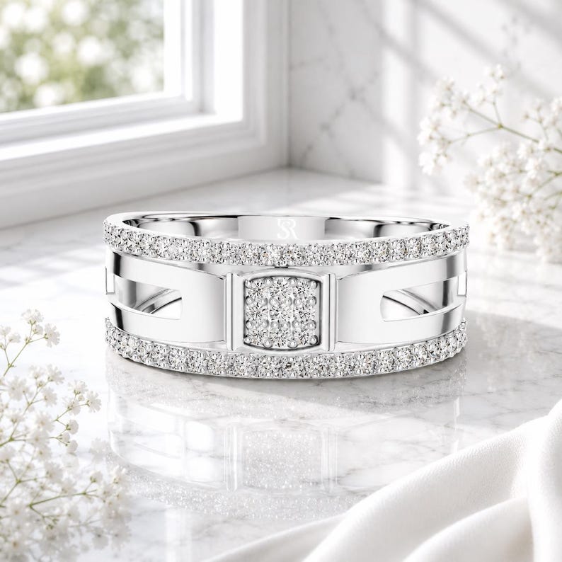 May include: A silver ring with a square center and a band of small, clear stones. The ring has a modern design with cut-out details and is set against a white background with flowers.