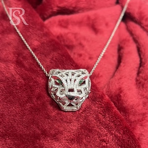Puede incluir: Un collar plateado con un colgante de cabeza de pantera. El colgante tiene un diseño calado detallado, con pequeñas piedras preciosas para los ojos. El collar se muestra sobre un fondo de tela roja.