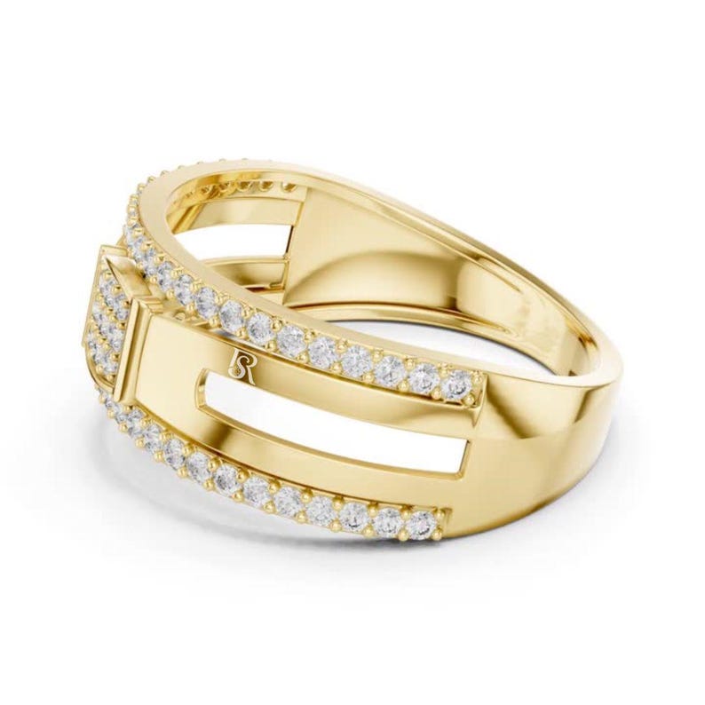 May include: A gold-colored ring with a rectangular design and a row of small, clear stones. The ring has a polished finish and a small letter "R" engraved on the side. The ring is set against a white background.