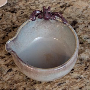 May include: A handmade ceramic bowl with a light blue interior and a dark purple rim. A small, stylized bat figure is perched on the rim. The bowl has a spout on one side.