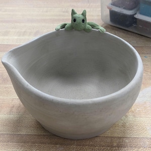 May include: A light gray ceramic bowl with a green cat figurine perched on the rim. The bowl is empty and has a smooth, slightly textured surface.